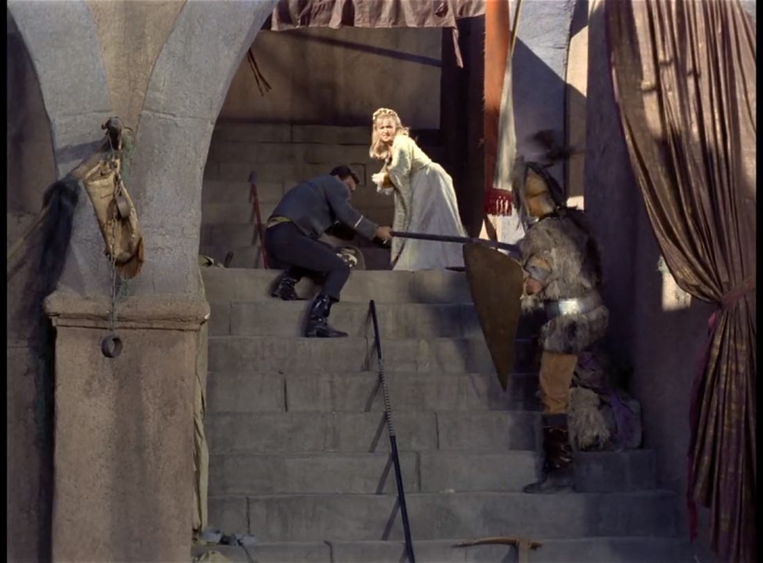 A set of stone stairs between an archway leading up to a balcony. A man in heavy furs and a helmet, carrying a shield, is advancing on Pike, who is crouched on the stairs holding him off with a spear. Behind Pike a woman in a white dress with long blonde hair is cowering