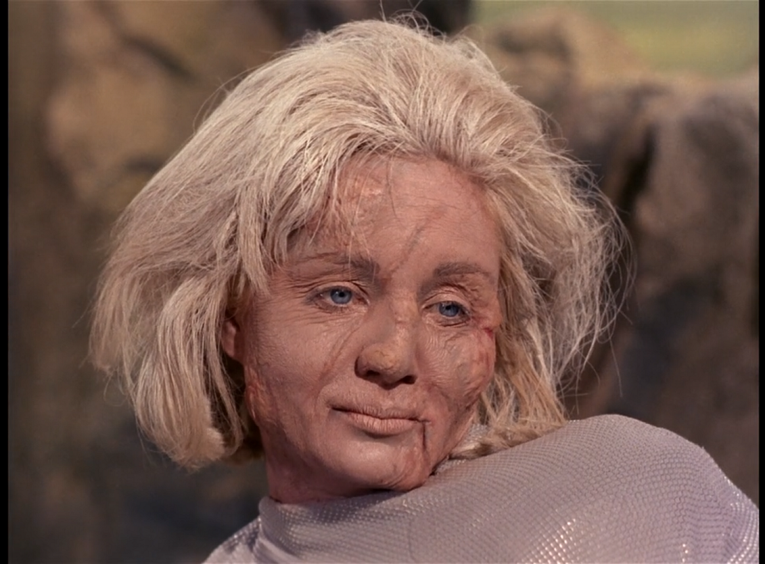 Vina, an old white woman with stiff pale hair, a scar running across her face and another under the corner of her mouth. Her right shoulder is hunched up close to her ear