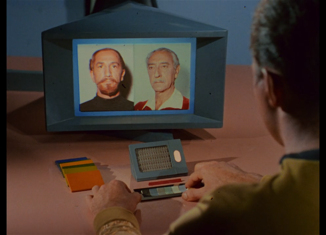 An over the shoulder shot of Kirk looking at a small computer screen on the table in front of him. The screen is showing two images side by side, one of a red-haired man with goatee, the other an older man with gray hair and a mustache.