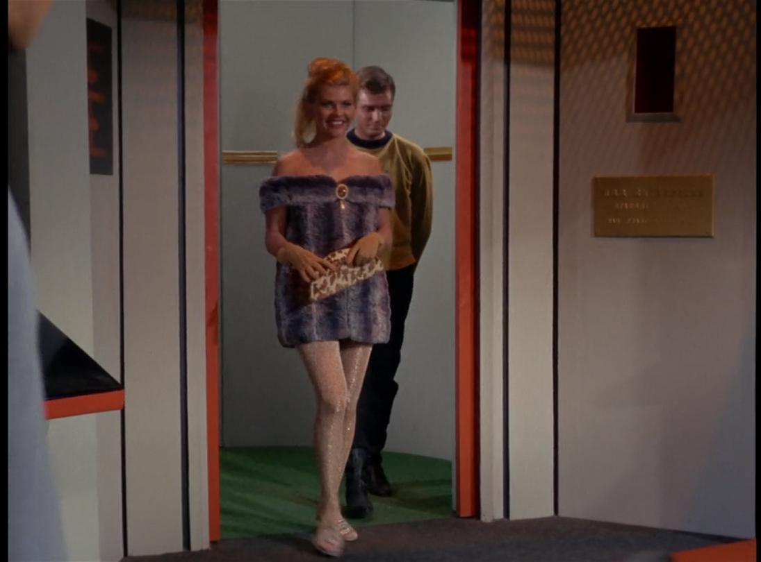 Lenore, a smiling white woman with blonde hair, walking through the doors of the turbolift to the bridge. She is wearing sparkly translucent tights and a furry gray dress-like thing with a boxy shape, which extends from partway down her arms to just below her hips. 