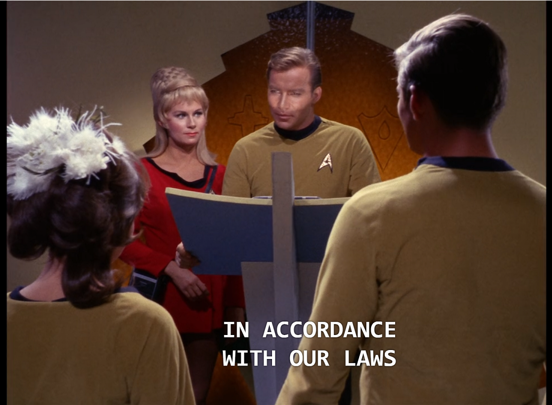 Kirk standing at a podium while Rand stands at his shoulder. Two goldshirts, a tall white man and a shorter white woman with flowers in her brown hair, are standing in the foreground. Kirk is saying, “In accordance with our laws...”