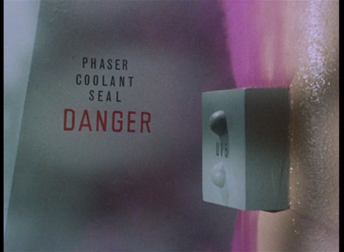 A gray box mounted on the wall, which is spewing pink gas from both ends. Text on the wall says ‘Phaser Coolant Seal DANGER’. 