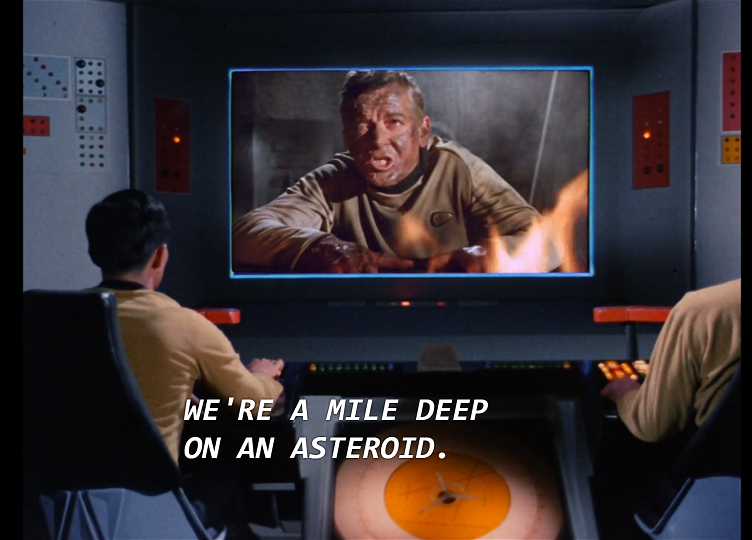 The bridge screen of the Enterprise, showing a white man in a tan uniform shirt, covered in dirt and sweat. There are flames in the foreground and dust and debris in the background. The man is saying, “We’re a mile deep on an asteroid.”