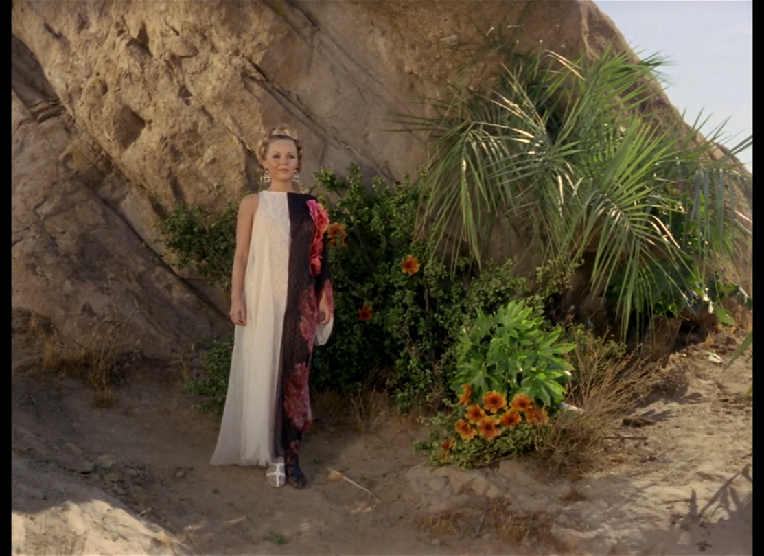 A white woman with blonde hair braided in a ring around her head, wearing a dress which is half white and half black with pink flowers, standing in front of a cliffside surrounded by plants.