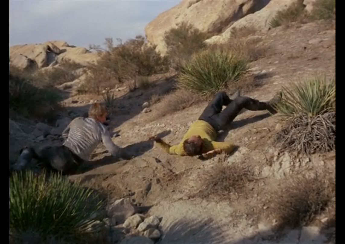 Kirk laying on his back on a rocky desert landscape, while Finnegan, a blonde-haired white man in a silver shirt, kneels on the ground nearby. 
