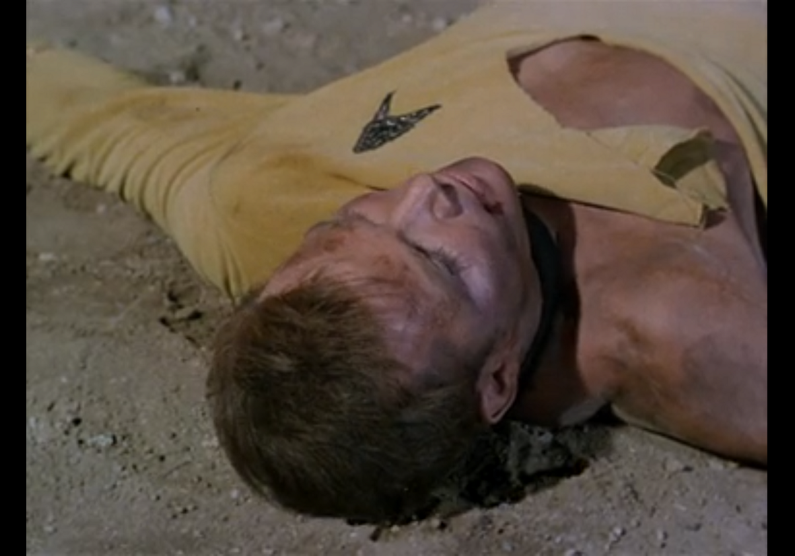 Kirk laying flat on his back, bruised and dirty, with his shirt torn off one shoulder almost to his stomach. 