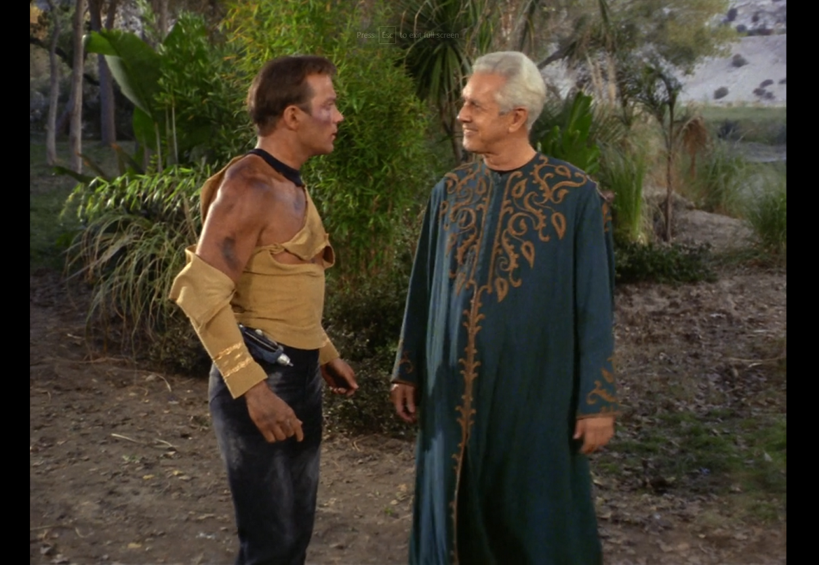 Kirk, still bruised with a badly torn shirt, looking in surprise at a kindly-looking white man with white hair, wearing a blue robe with gold leaf embroideries on the chest and cuffs.