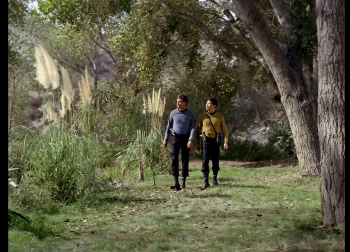 McCoy and Sulu walking down a sunlit grassy lane with trees to the right and tall plants at the edge of a pond to the left.