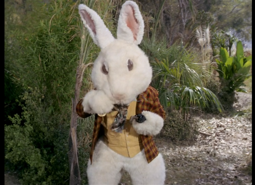A large humanoid white rabbit standing among the foliage, wearing a checked red and yellow shirt, yellow waistcoat, and brown and gray neckcloth, with an umbrella tucked under one arm.