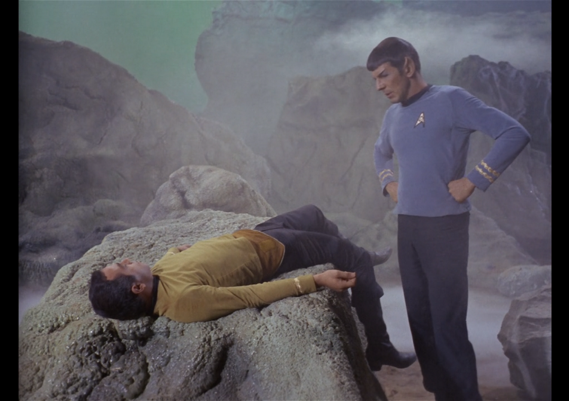  Gaetano’s dead body slumped over a rock while Spock stands over him with his hands on his hips.