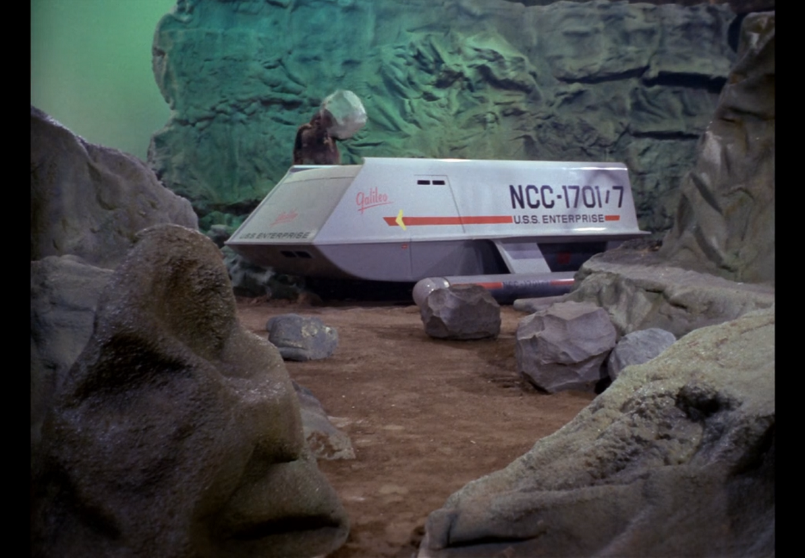 The shuttlecraft Galileo sitting among cliff walls while a giant stands over it with a large boulder poised to strike.