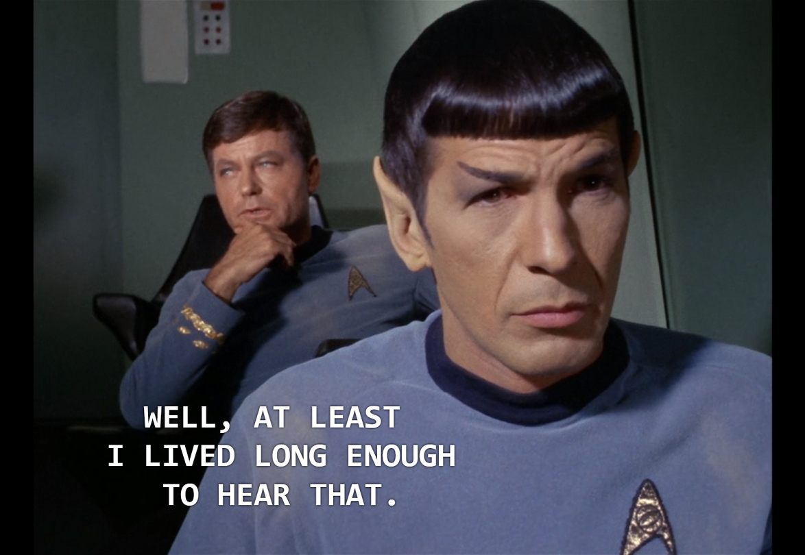 Spock sitting in the foreground while behind him McCoy, one hand on his chin, looks off to the side and says, “Well, at least I lived long enough to hear that.”