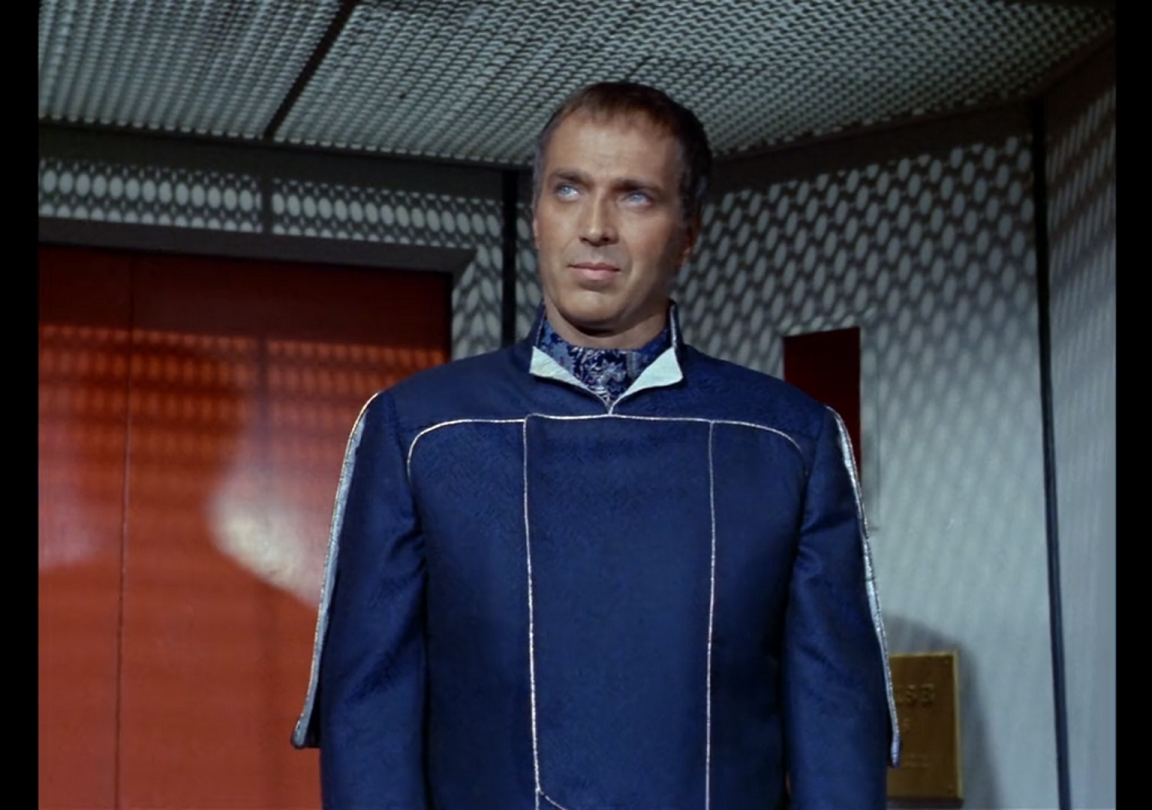 Ferris, a white man with very short graying brown hair, wearing a blue uniform with an elbow-length cape on the back.