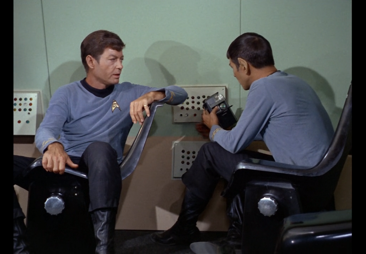 McCoy and Spock sitting inside the Galileo. McCoy is sitting sideways with his back to the wall and one arm propped against the back of the chair, looking over at Spock, who is sitting forward and looking at his tricorder.