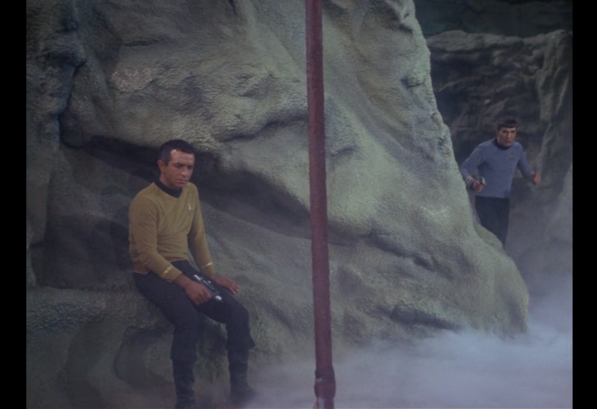 Gaetano, a white man with short dark hair, sitting on an outcropping in the side of a ravine and staring at a spearhaft that extends out of frame in both directions, while Spock approaches from around the corner.
