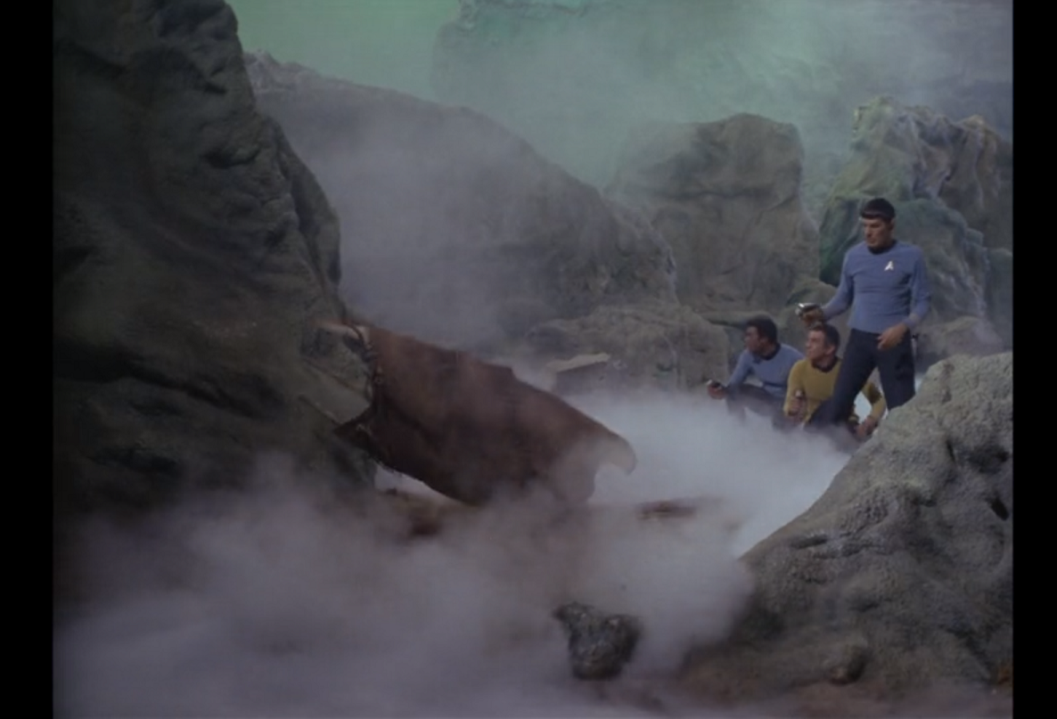 Spock stands in the ravine with his phaser out, Gaetano and Boma crouched at his side. A large, crude leather shield has fallen to the ground in front of them.