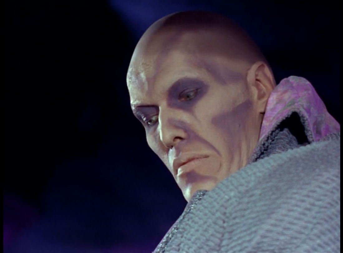 A bald white man with a face heavily contoured in purple, wearing a pale blue corduroy coat/robe thing with a dappled pink and green under-robe, looking dispassionately downward. 