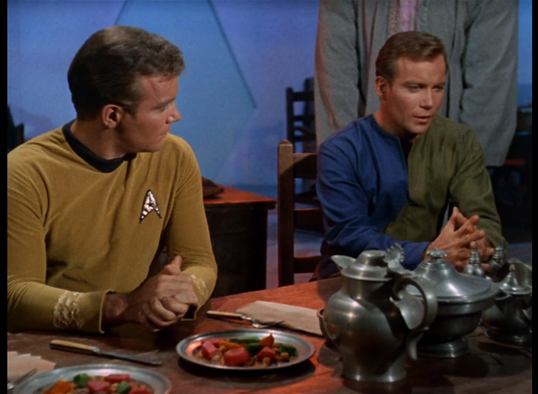 Two Kirks sitting side by side at a table with plates of food and covered dishes on it. One is wearing his standard gold uniform shirt while the other is wearing a blue and green jumpsuit. 