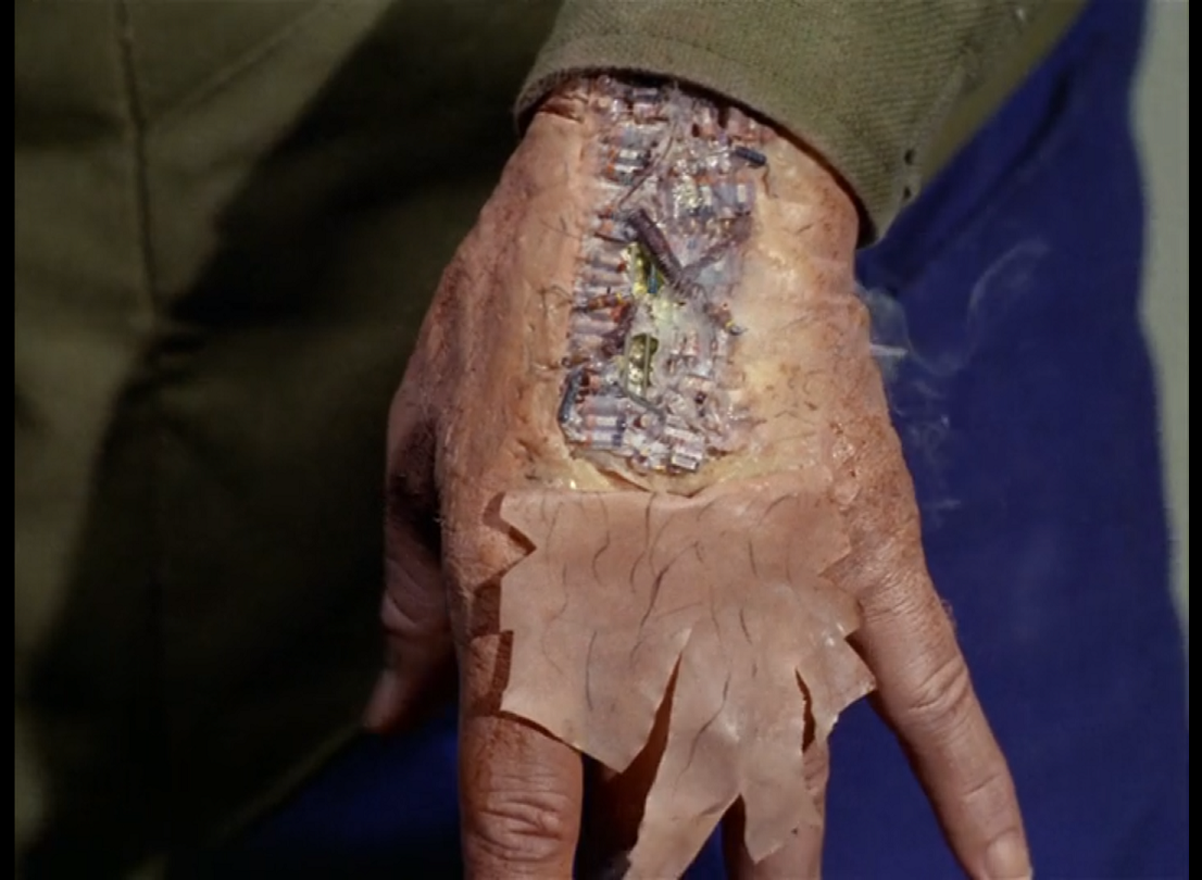 Korby's hand with the skin peeled off, showing a mess of smoking circuitry underneath. 