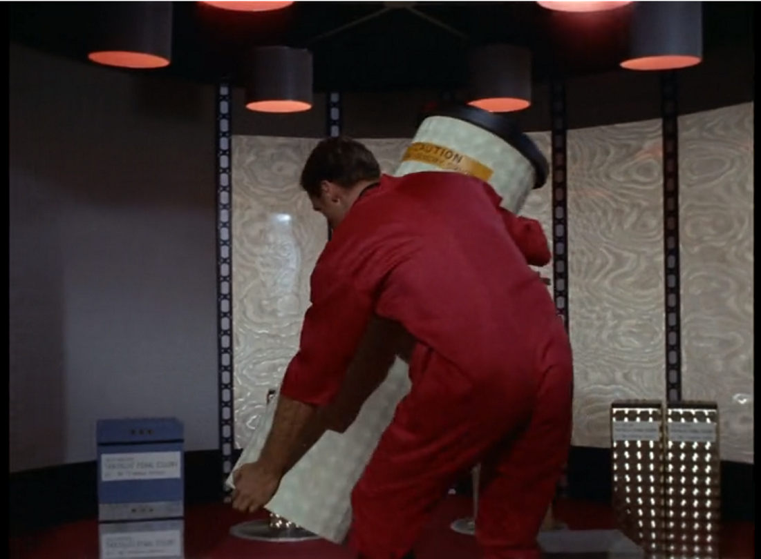 A red-jumpsuited crewman manhandling a giant white bottle onto the transporter pad. 