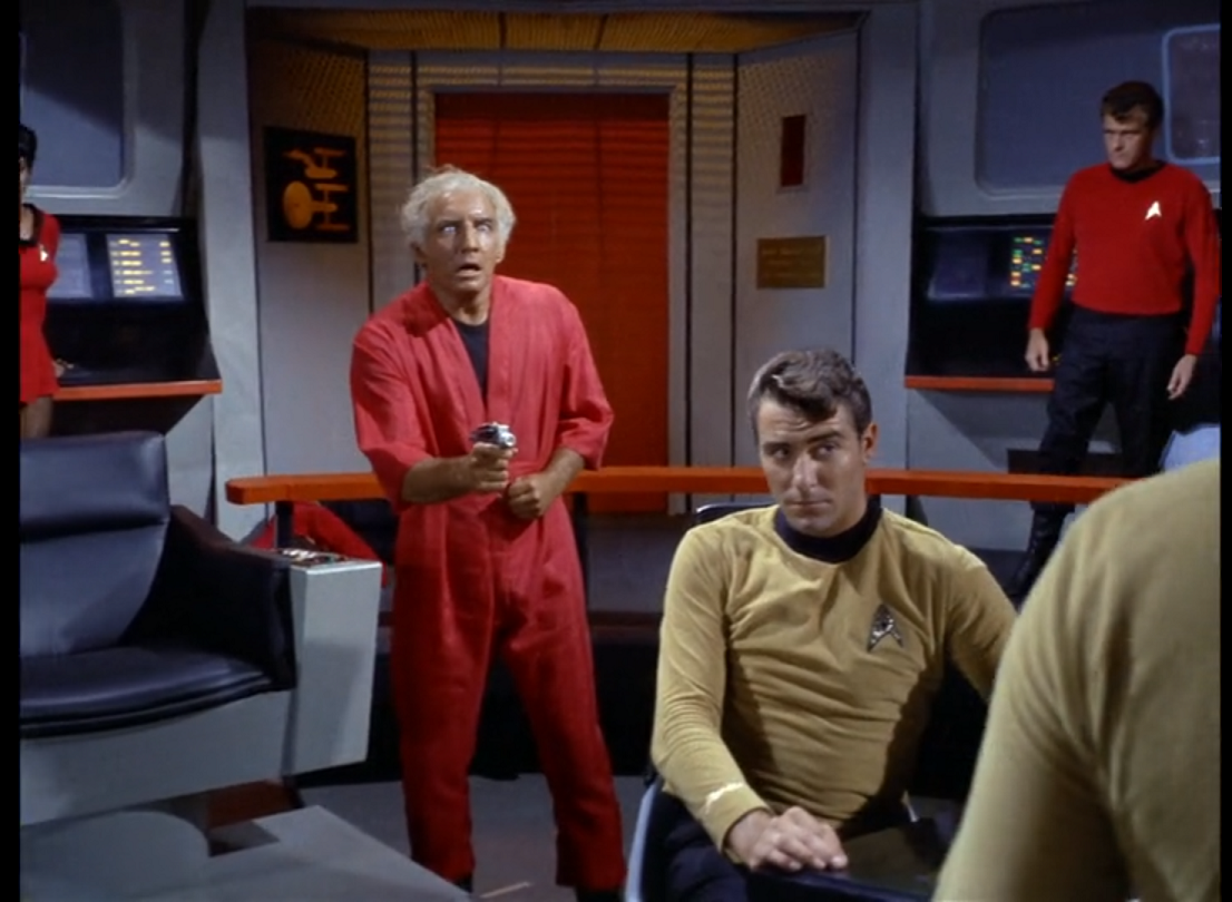 A scene on the bridge with Van Gelder, a white man with white hair and a wild expression, holding a phaser at Kirk, who is in the foreground. Between them is a goldshirt sitting at the helm station with a less than impressed expression on his face. 