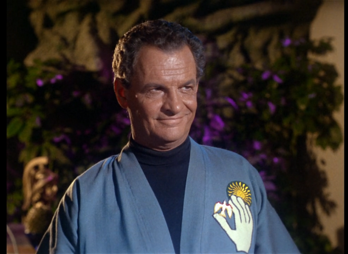 A middle-aged white man with short graying brown hair, wearing an open blue robe-shirt over a dark blue turtleneck, standing in front of a background of flowers and plants. The man’s shirt has a patch on the left breast showing a white hand holding a dove against a stylized sun. He has quite a smug smirk on his face.